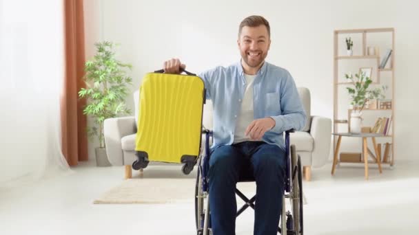 Feliz persona discapacitada en una silla de ruedas con una maleta amarilla en sus manos se prepara para un viaje — Vídeo de stock