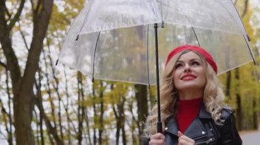 Kırmızı elbiseli romantik sarışın ve parkta duran şeffaf şemsiyeli motosiklet ceketli.