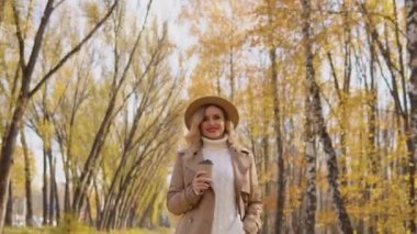 Kahverengi paltolu ve şapkalı genç bir kadın sonbahar parkında yürüyor ve kahve içiyor.