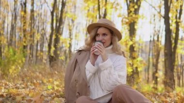 Kahverengi şapkalı güzel sarışın kadın sonbahar parkında kahve içiyor.