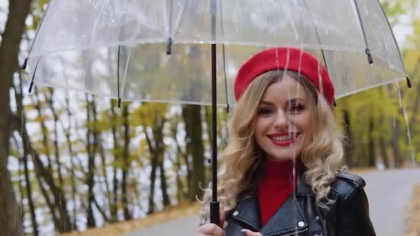 Sorrindo mulher alegre feliz em um terno vermelho e uma jaqueta de motociclista com um guarda-chuva transparente em um dia chuvoso. Doenças sazonais, tosse, frio — Vídeo de Stock