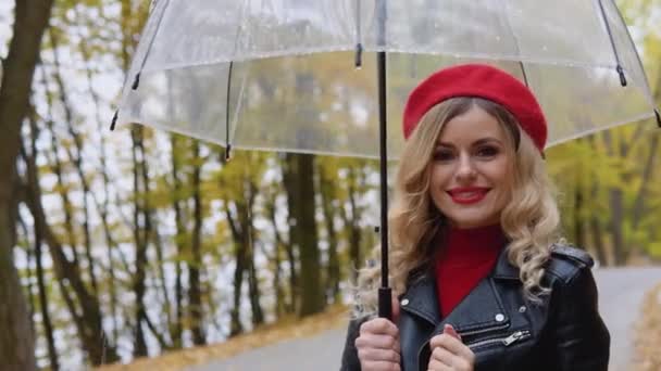 Mosolygó boldog, vidám nő piros ruhában és egy bringás kabát átlátszó esernyővel egy esős napon — Stock videók