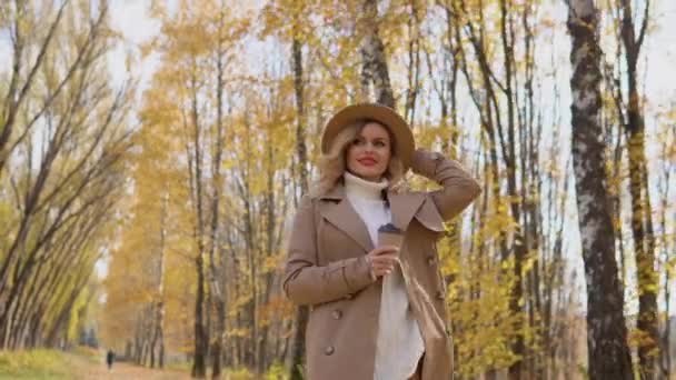 Молодая женщина в коричневом пальто и шляпе гуляет по осеннему парку и пьет кофе — стоковое видео