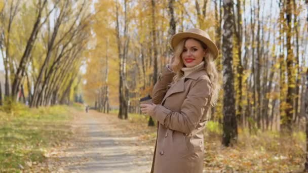 Молодая женщина в коричневом пальто и шляпе гуляет по осеннему парку — стоковое видео