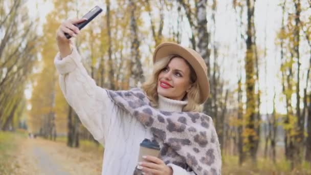 Beautiful blonde woman in a brown hat takes a selfie in the autumn park — Stock Video