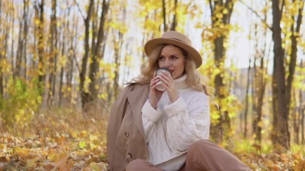 Kahverengi şapkalı güzel sarışın kadın sonbahar parkında kahve içiyor. — Stok video
