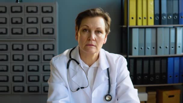 Doctora mayor vistiendo bata médica blanca y estetoscopio mirando la cámara. Médico femenino posando en la oficina del hospital — Vídeos de Stock
