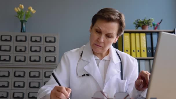 シニア女性医師50は、ラップトップで動作し、患者の医療史を書き込みます — ストック動画
