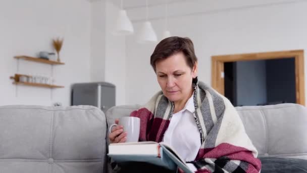 Seniorin liest ein Buch, während sie zu Hause auf der Couch sitzt. Lebensstil der selbstisolierten Frau in Quarantäne — Stockvideo