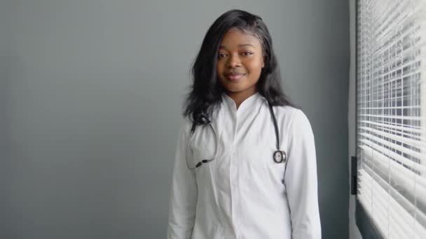 Beyaz önlüklü ve steteskoplu yakışıklı Afrikalı kadın doktorun portresi, sağlık kliniğinde duruyor. — Stok video