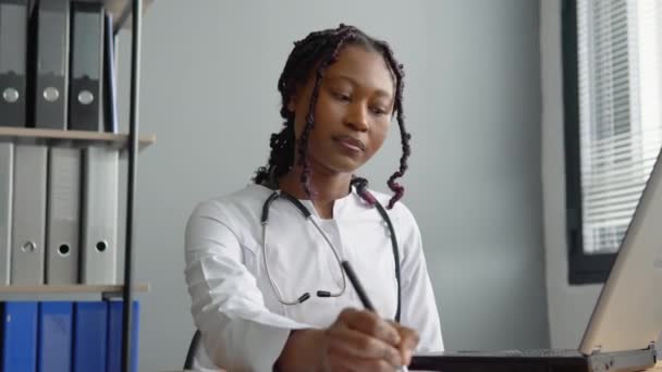 Mujer afroamericana joven médico trabaja con un ordenador portátil y escribe la historia clínica de los pacientes — Vídeos de Stock