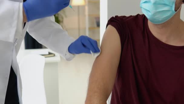 Ο γιατρός που χορηγεί το εμβόλιο Covid-19 ή το εμβόλιο κατά του ιού της γρίπης σε νεαρό άνδρα ασθενή φοράει μάσκα προστασίας του προσώπου από την ιογενή νόσο σε κλινική υγείας ή νοσοκομειακό γραφείο. Έννοια εμβολιασμού — Αρχείο Βίντεο