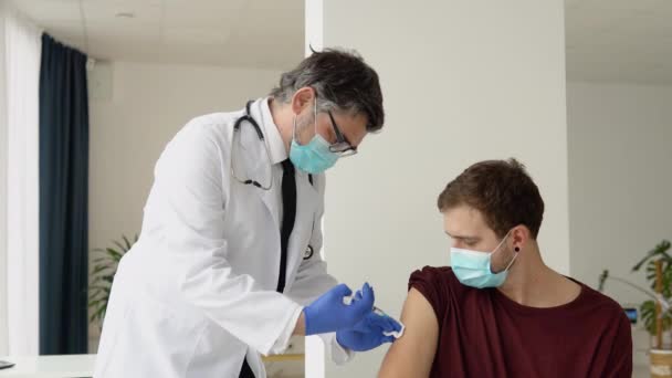 Starší lékař podávající Covid-19 nebo vakcínu proti viru chřipky mladým mužským pacientům nosí na klinice nebo v nemocničním úřadě ochranu před virovou chorobou. Koncepce očkování — Stock video