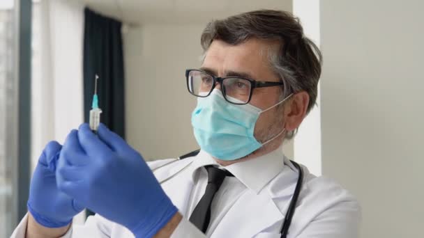 Старший врач-мужчина готовит шприц для вакцинации. В маске для лица и хирургических перчатках. Вакцина Ковида-19 — стоковое видео