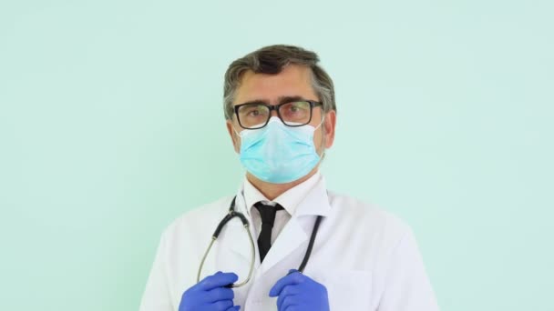 Portrét staršího lékaře v bílém obleku a ochranné masky na modrém pozadí — Stock video