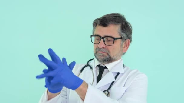 Oberarzt mit Schutzmaske zieht medizinische Handschuhe auf blauem Hintergrund an — Stockvideo