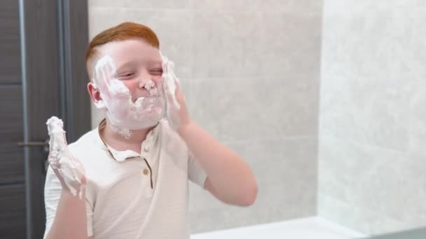 Chico divertido en el baño unta su cara con crema de afeitar, el niño imita a un padre adulto afeita — Vídeos de Stock