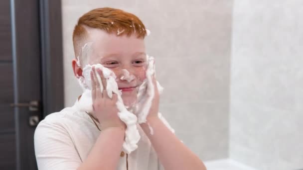 Chico divertido en el baño unta su cara con crema de afeitar, el niño imita a un padre adulto afeita — Vídeos de Stock