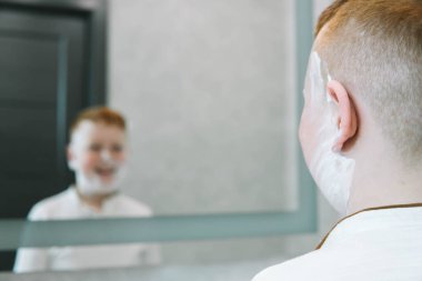 Banyodaki komik çocuk yüzünü tıraş köpüğüyle boyuyor, çocuk da yetişkin bir babayı taklit ediyor. Çocuk tıraş köpüğü ile yanaklarını lekeledi.