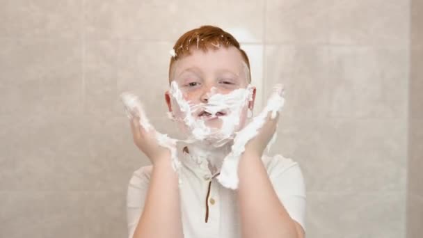 風呂で面白い少年はシェービングクリームで顔を笑顔、子供は大人のお父さんのシェービングを模倣します。男の子は髭を剃りながら頬をしかめた。 — ストック動画