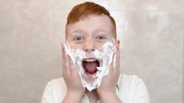 Pojken smetade in sina kinder med raklödder. Rolig pojke i badet smetar sitt ansikte med rakkräm, barnet imiterar en vuxen pappa rakar — Stockvideo