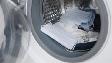 Erkek el, giysileri ve tıbbi maskeleri çamaşır makinesine koyar ve kapıyı kapatır. Çamaşır makinesi yükleniyor