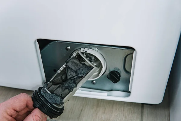 La mano masculina elimina el filtro de bomba de lavadora sucia y obstruida. Reparación de electrodomésticos en casa con sus propias manos. el reparador repara la lavadora Imagen De Stock