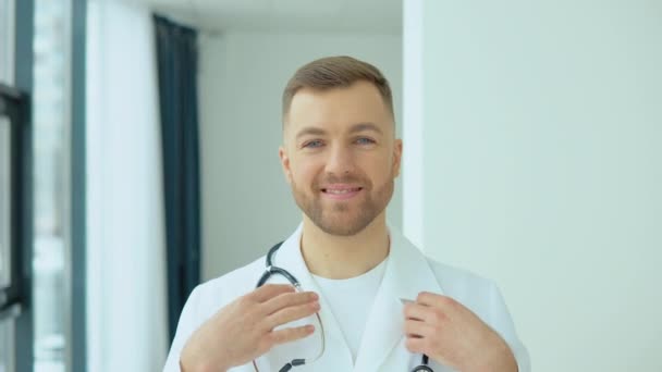 Udany lekarz w białym fartuchu patrzy w kamerę i uśmiecha się w gabinecie szpitalnym. — Wideo stockowe