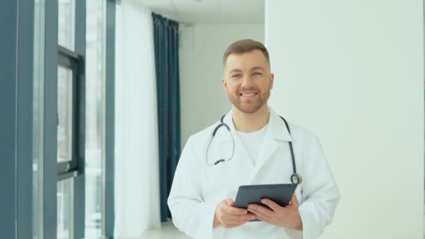 Успішний лікар у білому лабораторному пальто з планшетом дивиться на камеру і посміхається в лікарняному кабінеті — стокове відео