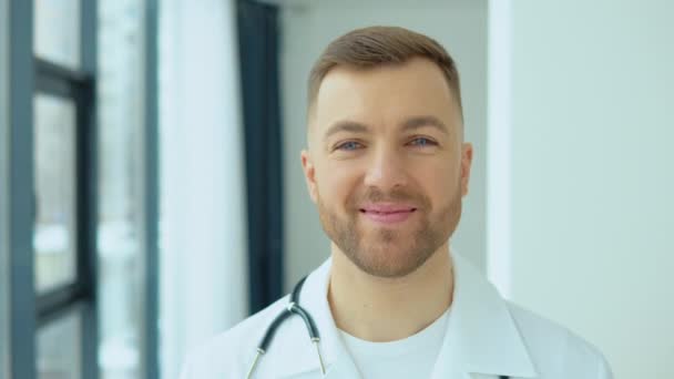 Udany lekarz w białym fartuchu patrzy w kamerę i uśmiecha się w gabinecie szpitalnym. — Wideo stockowe