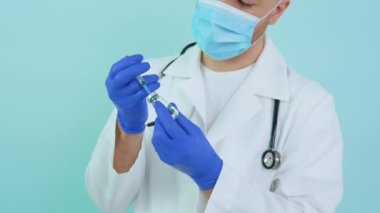 Koruyucu maskeli genç bir doktor mavi arka planda bir şırıngaya aşı çiziyor.