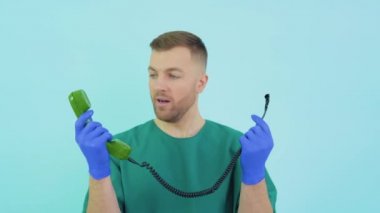 Yeşil üniformalı ve mavi eldivenli doktor eski bir telefonu elinde tutarken şaşırır.