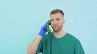 Yeşil üniformalı ve mavi eldivenli bir doktor eski bir telefonla konuşuyor ve mavi arka plandaki bir hastaya danışıyor.