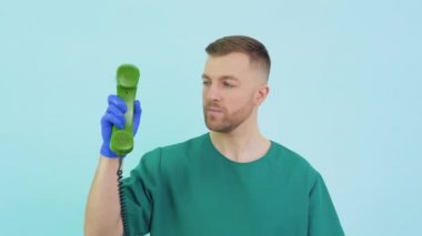 Yeşil üniformalı ve mavi eldivenli doktor eski bir telefonu elinde tutuyor ve mavi arka planda onu işaret ediyor.