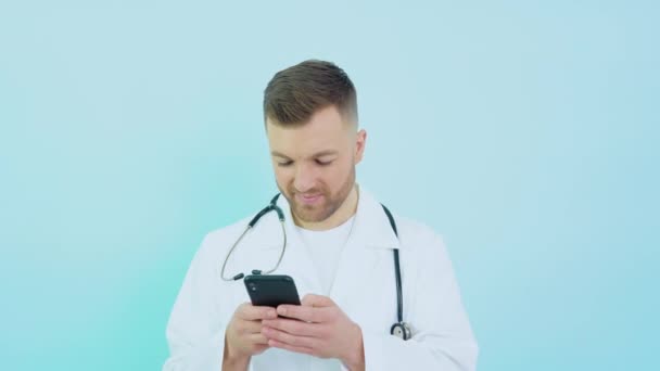Doctor con estetoscopio en bata blanca se corresponde en un teléfono inteligente sobre un fondo azul — Vídeos de Stock