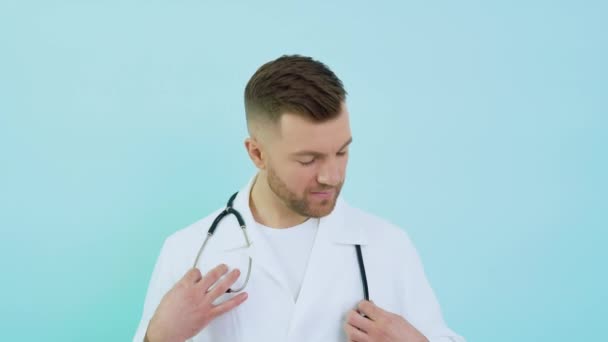 Successful physician in white lab coat looks at the camera and smiles on a blue background — Stock Video