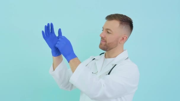 Młody lekarz w masce ochronnej zakłada rękawiczki medyczne na niebieskim tle — Wideo stockowe