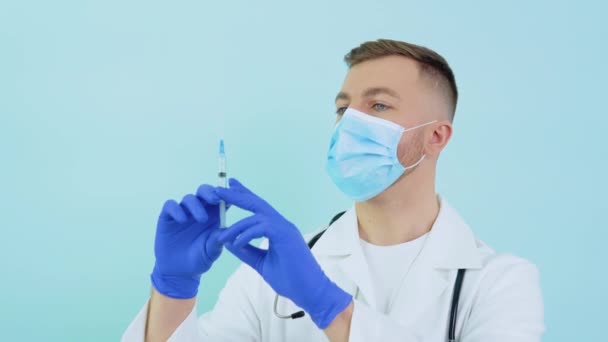 Männlicher Arzt in Schutzmaske mit einer Impfspritze in den Händen auf blauem Hintergrund — Stockvideo