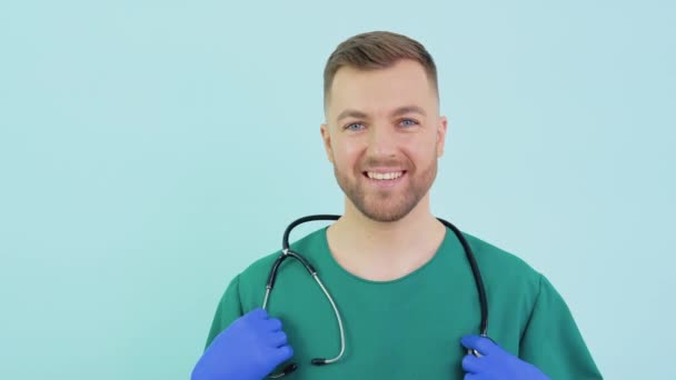 녹색 제복에 푸른 장갑 과 청진기를 착용 한 성공적 인 의사는 카메라를 보고 파란 배경에 미소를 짓습니다 — 비디오
