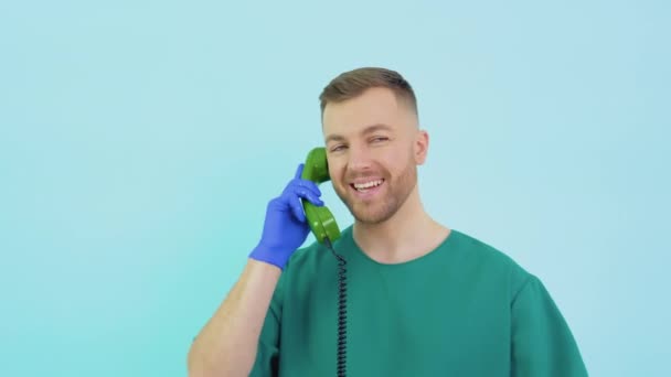 A friendly doctor in a green uniform and blue gloves speaks on an old telephone and consults a patient — Vídeos de Stock