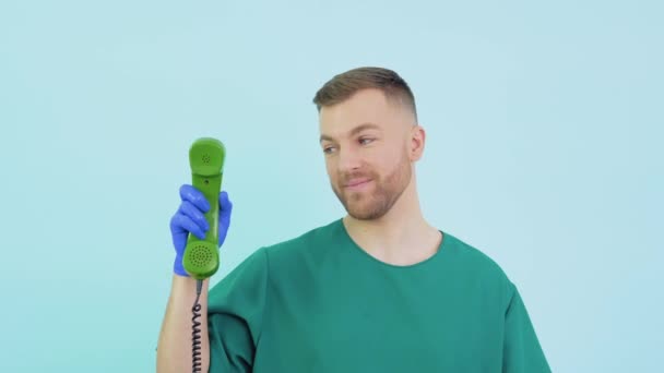 Um médico amigável com um uniforme verde e luvas azuis segura um telefone antigo em suas mãos e aponta para ele — Vídeo de Stock