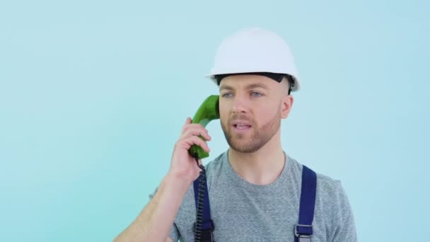 Serviceman in overalls and helmet speaks on an old telephone and consults a client on a blue background — Stock Video