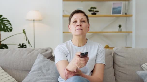 Signora anziana guardando la tv in soggiorno seduto sul divano in possesso di telecomando cambiando i canali televisivi. Donna anziana con telecomando TV — Video Stock