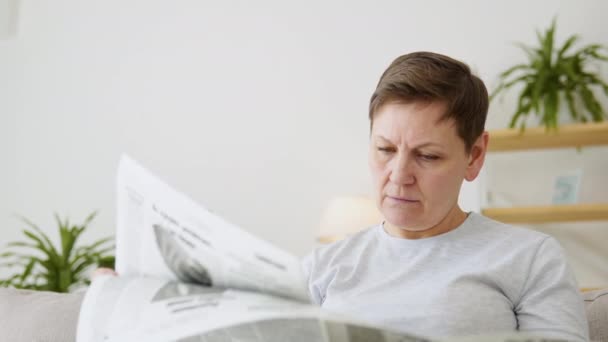 A close-up view of a beautiful mature woman is reading a newspaper sitting on sofa at home in the morning — Stock Video