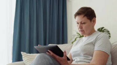 Kanepede yalnız başına dijital tablet kullanan gülümseyen olgun bir kadın. Mutlu emekliler web sörfü bilgileri. İnternetten öğrenim ya da alışveriş