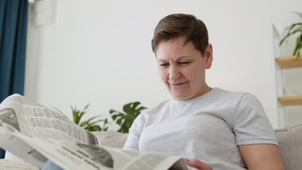 Una visione ravvicinata di una bella donna matura sta leggendo un giornale seduto sul divano a casa al mattino. Ritratto di donna anziana che legge il giornale — Video Stock