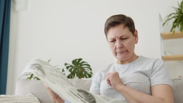 A close-up view of a beautiful mature woman is reading a newspaper sitting on sofa at home in the morning — Stock Video