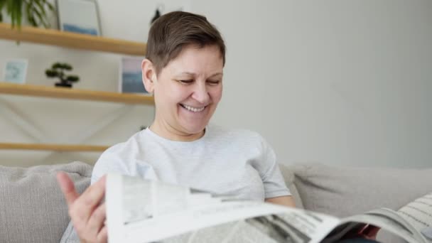 En närbild av en vacker mogen kvinna läser en tidning som sitter på soffan hemma på morgonen. Porträtt av äldre kvinna läser tidningen — Stockvideo