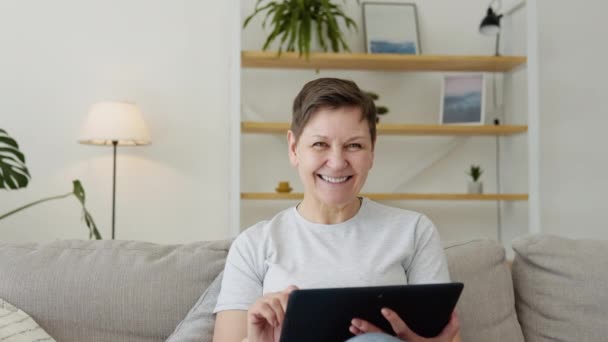 Sorridente anziana donna matura che riposa sul divano, utilizzando tablet digitale da solo a casa — Video Stock