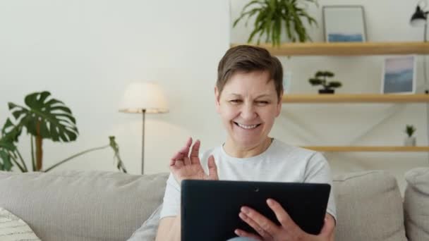 Kanepede yalnız başına dijital tablet kullanan gülümseyen olgun bir kadın. Sosyal ağlarda sohbet. — Stok video
